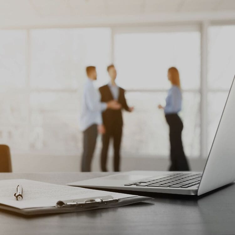 Schreibtisch mit Laptop und Notizblock, im Hintergrund ein IT-Projektmeeting in modernem Büro